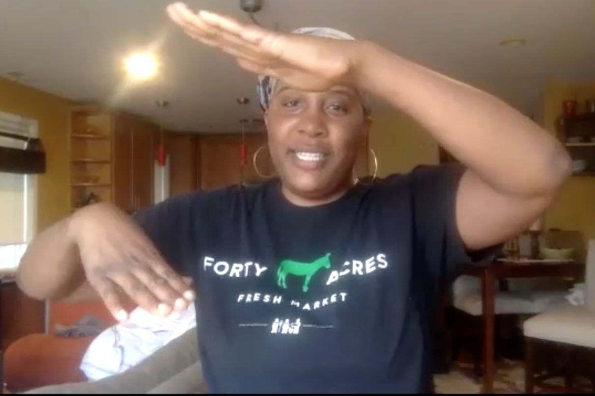 Screenshot of Liz Abunaw from a Zoom call with hands and arms gesturing across her body. She is wearing a shirt with forty acres fresh market logo.