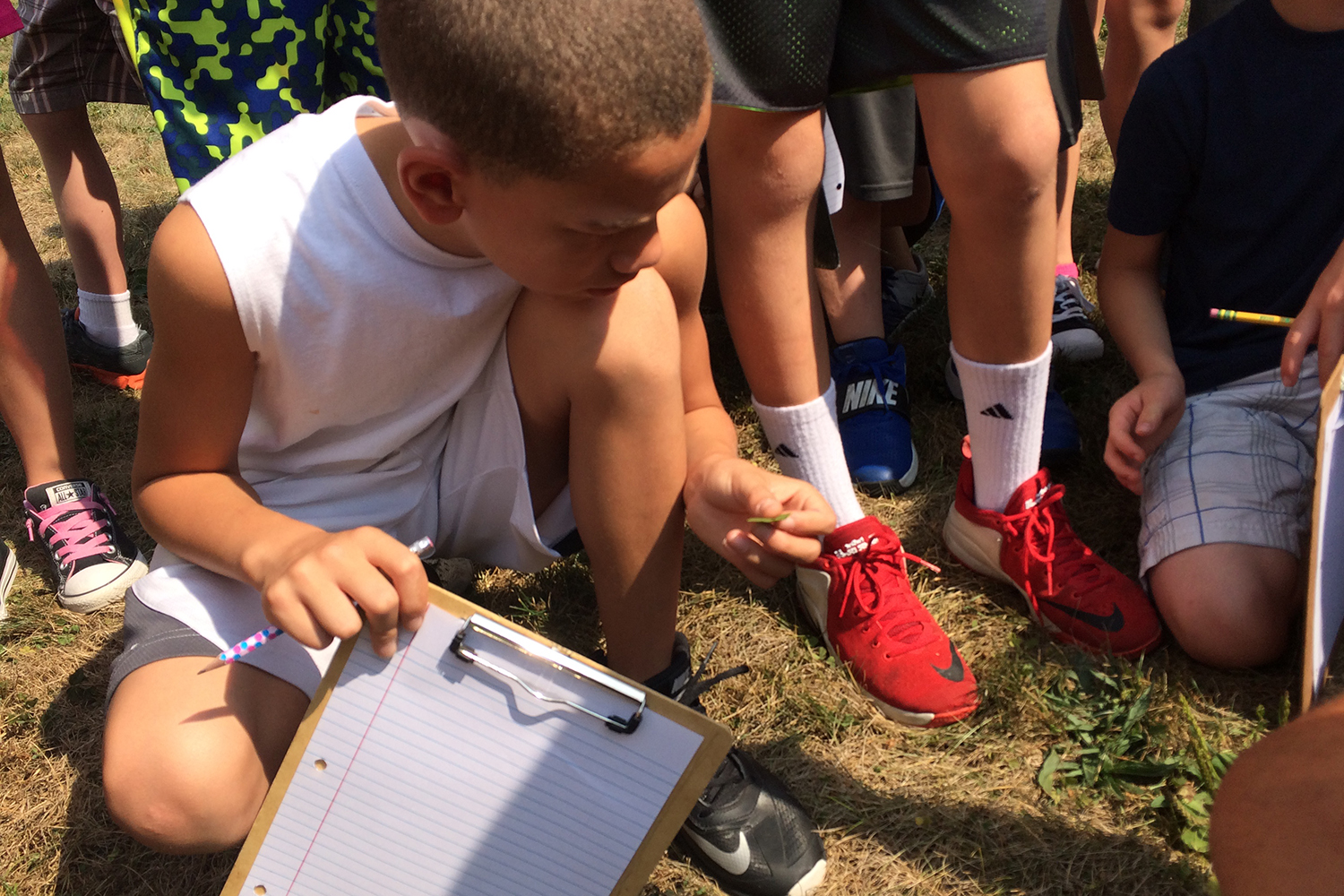 A kid squatting down on the ground, on knee up, examining a leaf, holding a clipboard and pencil, lots of shoes and legs of other kids in the background.