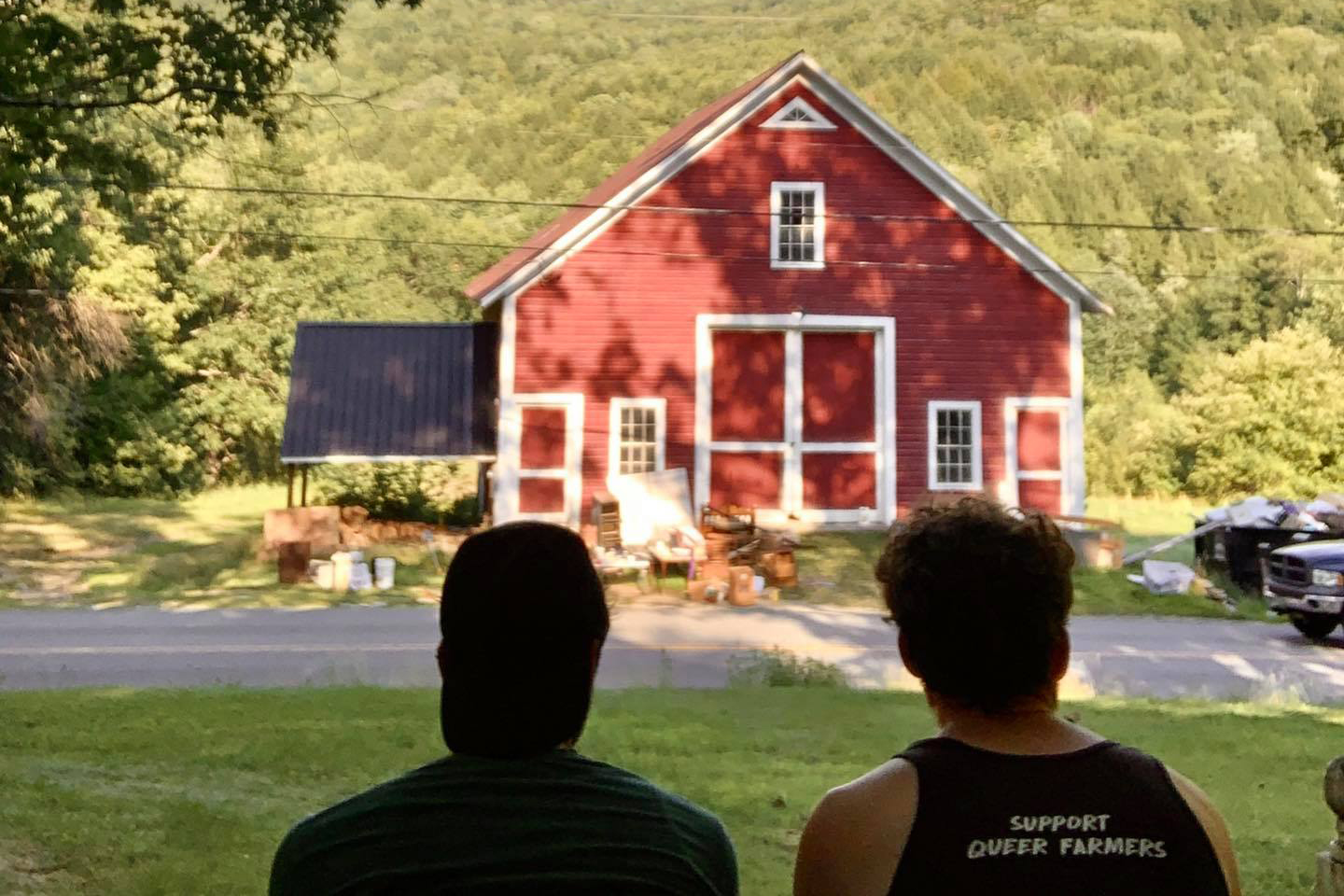 Two figures from the back, looking at a red barn. One of the figures has 'support queer farmers' on the back of their shirt