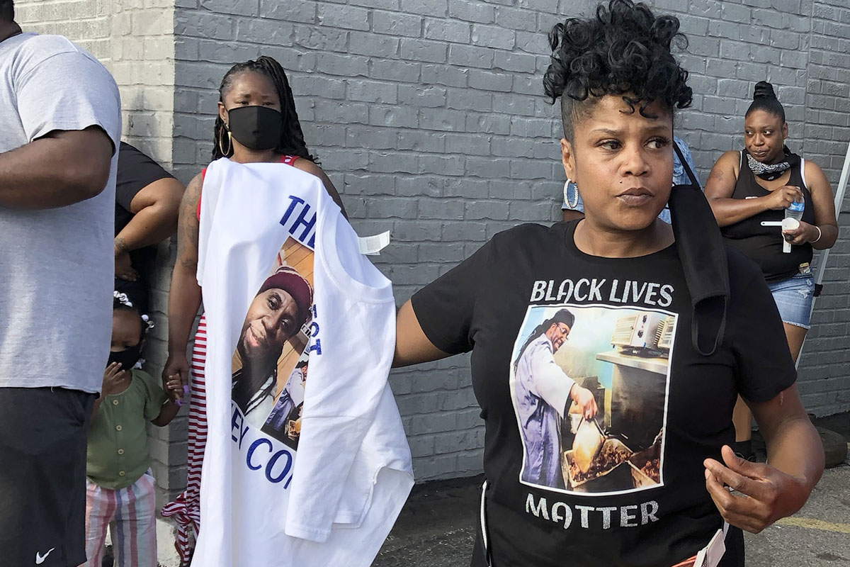 Marcia Lawhorn, with black face mask hanging from one ear, wearing t-shirt with David McAtee, cooking, and "Black Lives Matter" holding up another shirt, people in facemasks in the background.