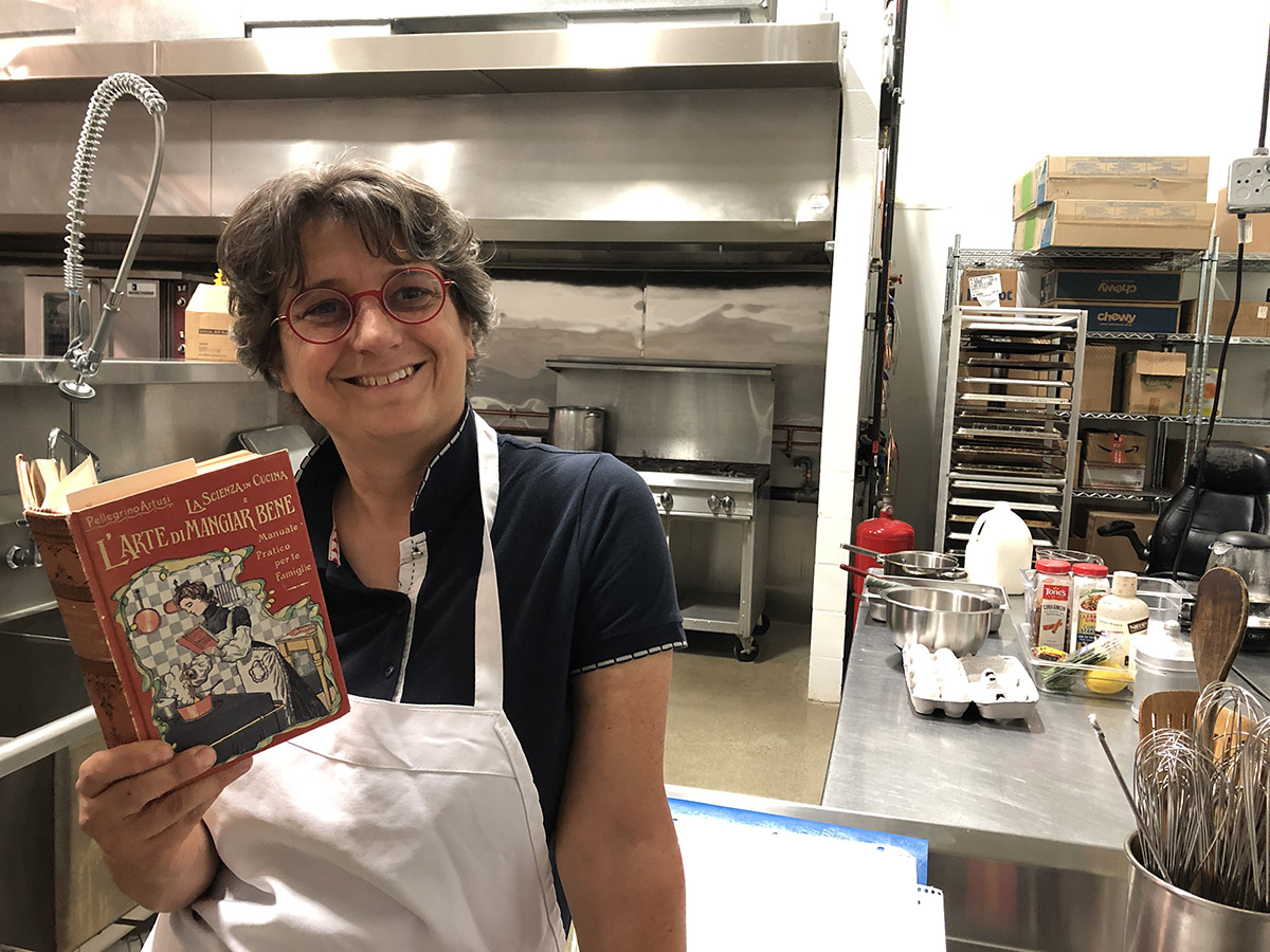 Maria Carlassare standing in a commercial kitchen holding a book