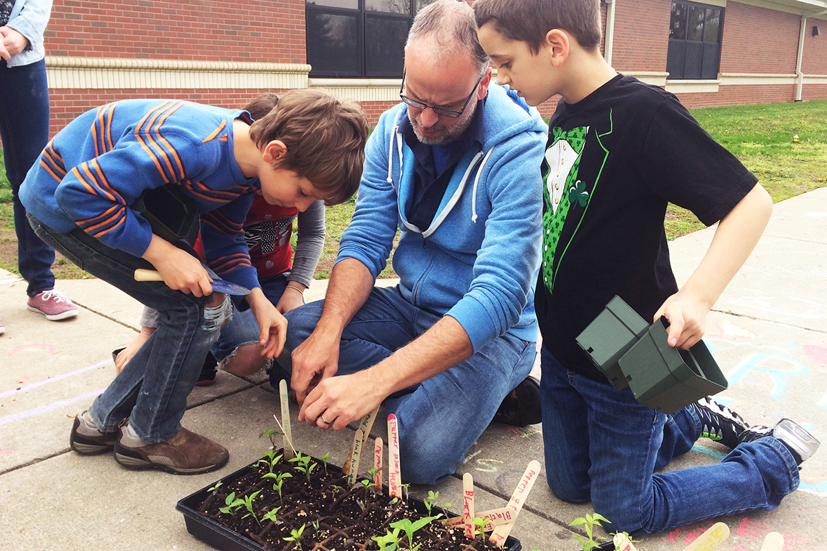 Mark Minster on the ground with a tray of seedlings, kids surrounding him,