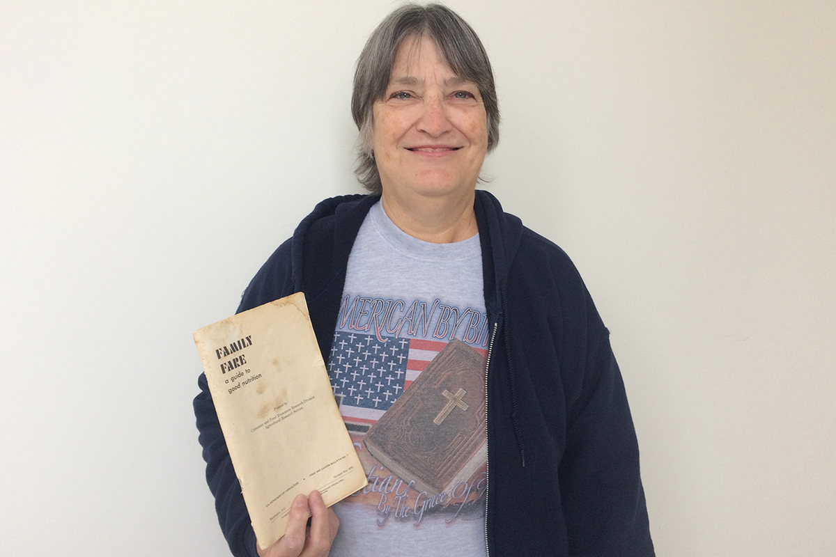Michelle Porter standing holding a yellowing booklet called Family Fare. She stands in front of a plain white background, wearing a t-shirt with American flag and Bible.  