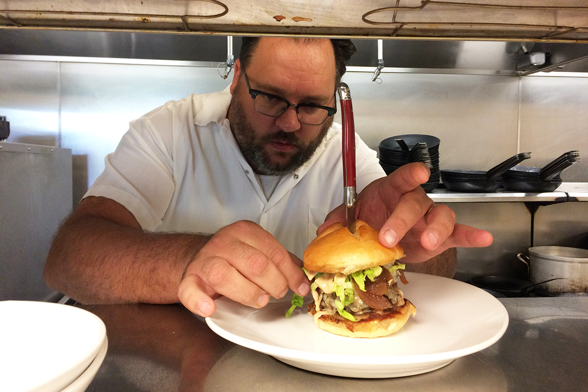 Ryan Nelson with a burger on a plate with a knife in the top, restaurant kitchen in background