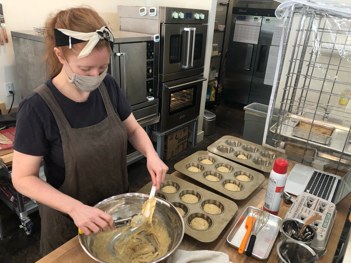 Nicole Evans Groth standing in a commercial kitchen with face covering and scarf holding back her hair. She is scooping batter from a steel bowl, muffin tins with batter are next to the bowl.