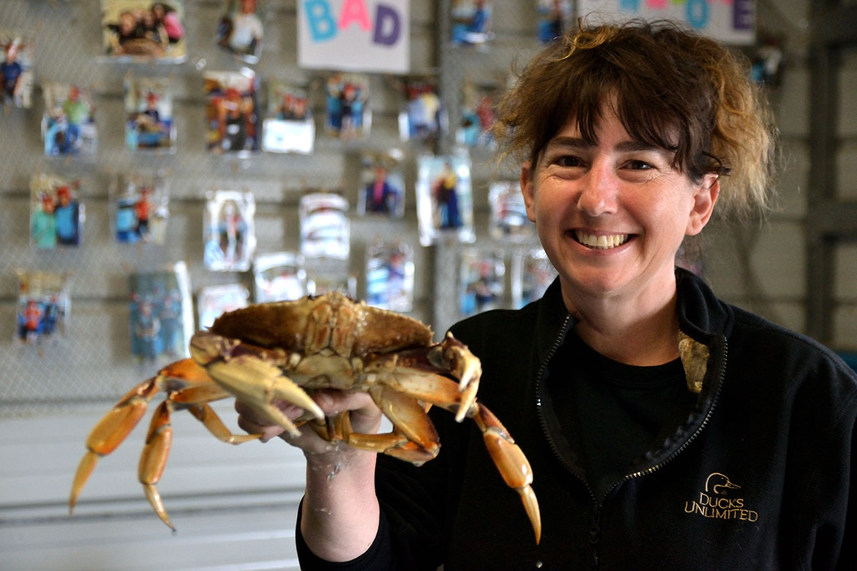 Amber Novelli smiling and holding a dungeness crab at shoulder height, indoors.