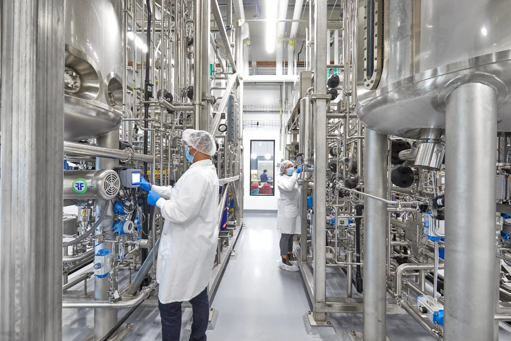 Two people with white coats in a hall surrounded by steel pipes