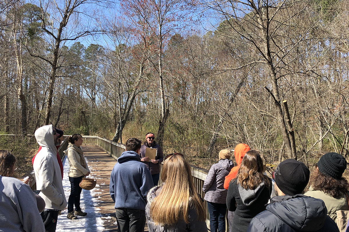 People gathered around a woman outdoors on a snowy boardwalk with bare trees and blue skies in the background.