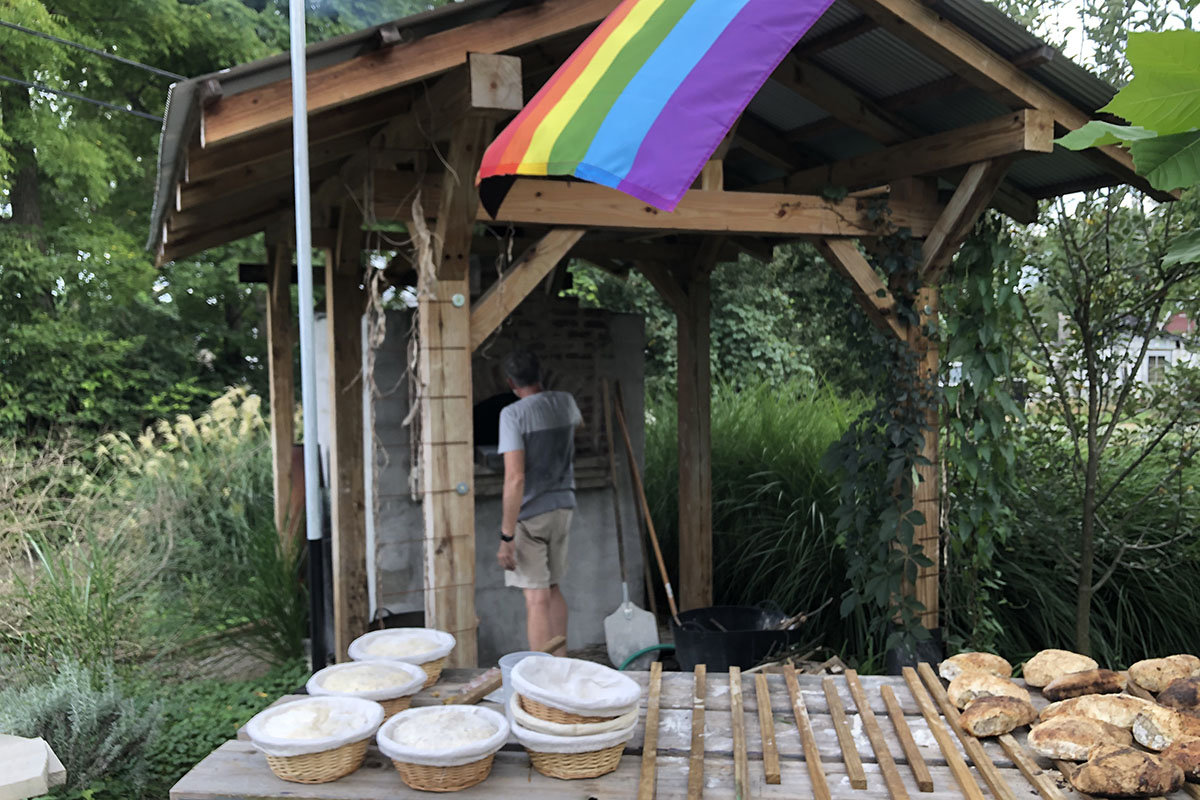 View of a person in front of a brick oven, with a small roof, a rainbow flag and a table with baked bread and dough on a long table, outside in a patio space surrounded by plants and trees.