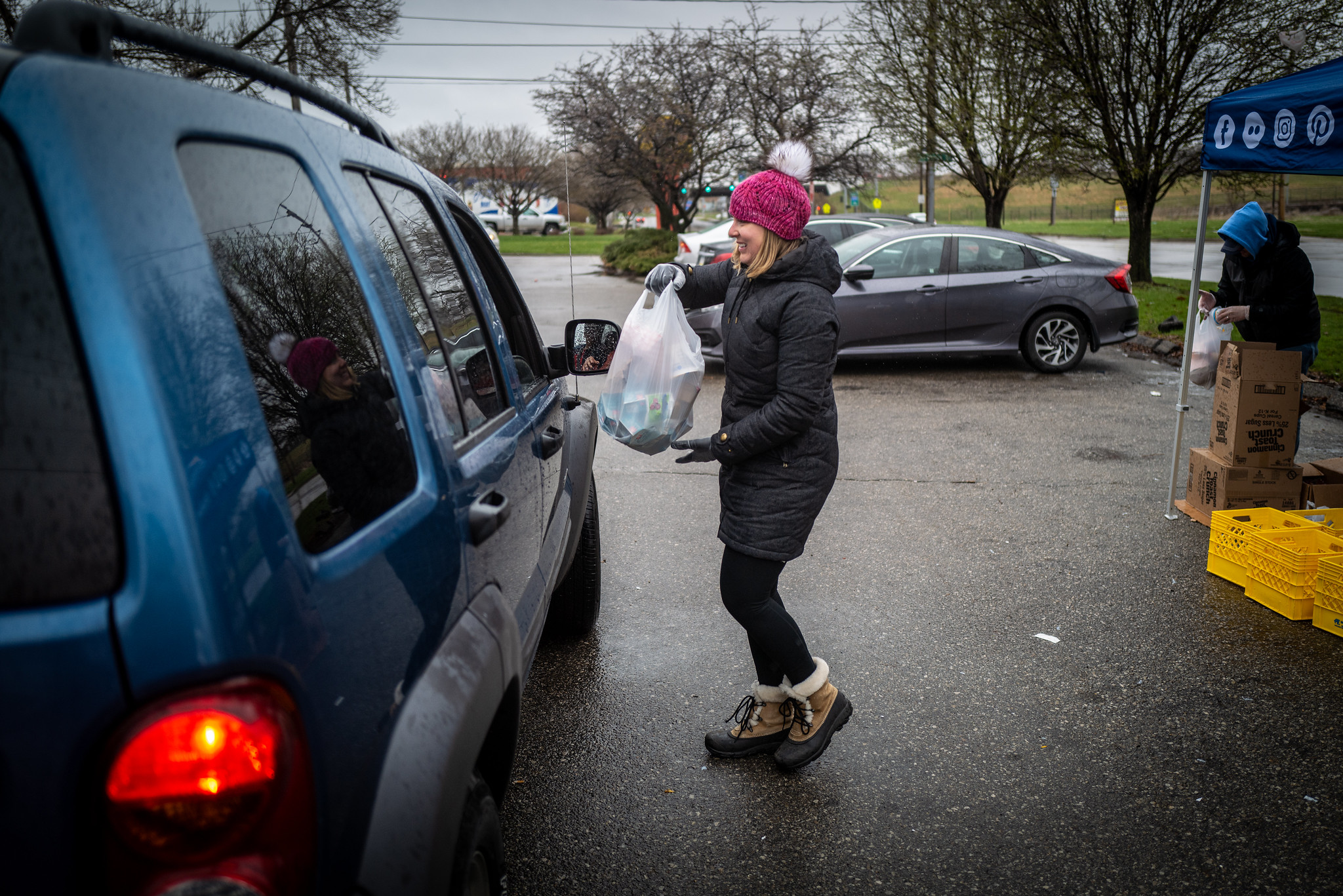 A smiling blonde woman in coat, hat and gloves approaches a car window with a full plastic bag