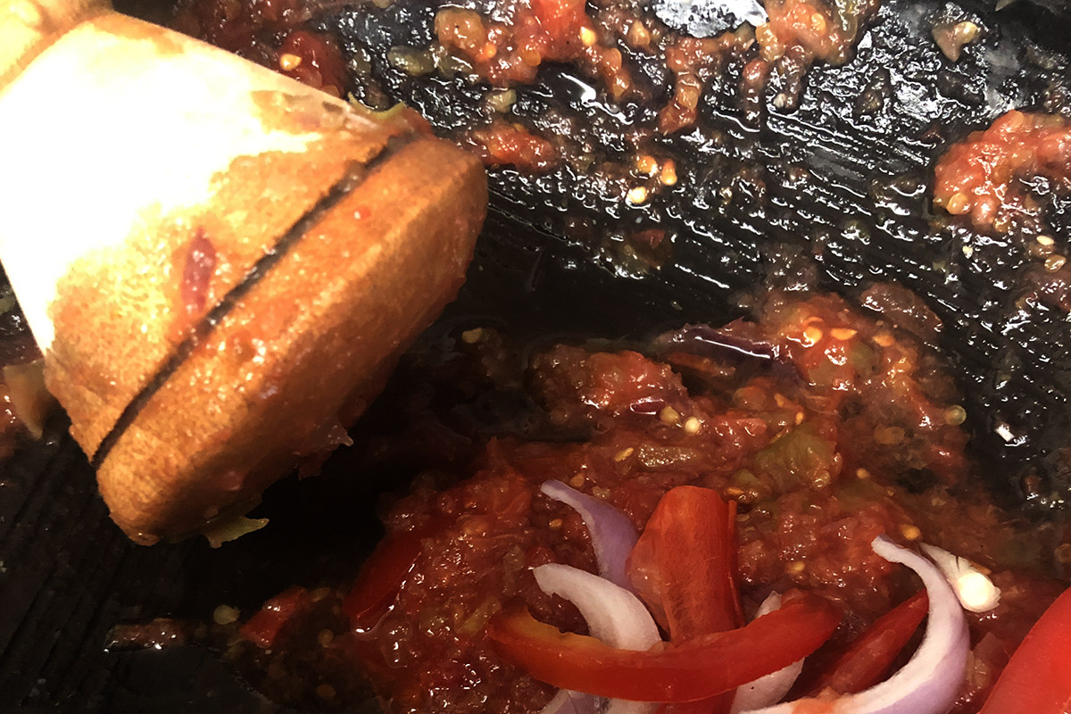 Close up of crushed tomato sauce in black pan with wooden pestle and onion and pepper slices.