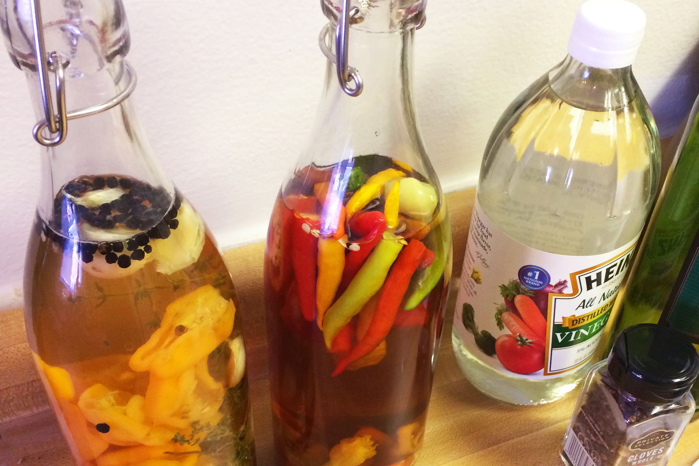 Two bottles of liquid with peppers and spices on a counter, with vinegar bottle and pepper corns bottle. 