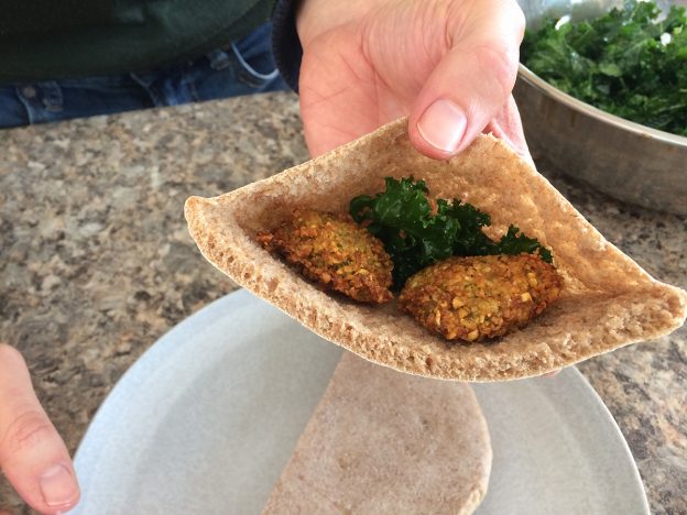 A hand holding a pita pocket open, inside is falafel and some dark, leafy greens.