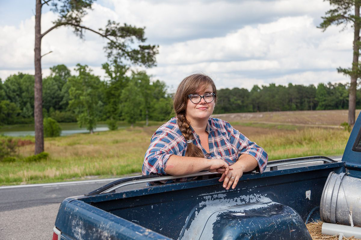 Rachel Herrick stands in plaid shirt with arms draped over the bed of a weathered pickup truck with trees lake and field in the background