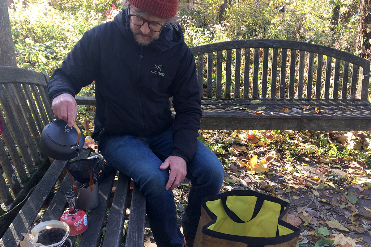 River Bailey sitting on a bench pouring water to make coffee outide.