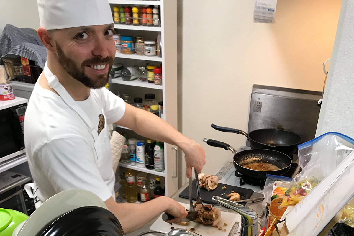 Rocky Burton, cutting something that looks like meat, turning towards the camera in the middle of a small kitchen with pots and pans all around, and an orderly spice rack in the background.