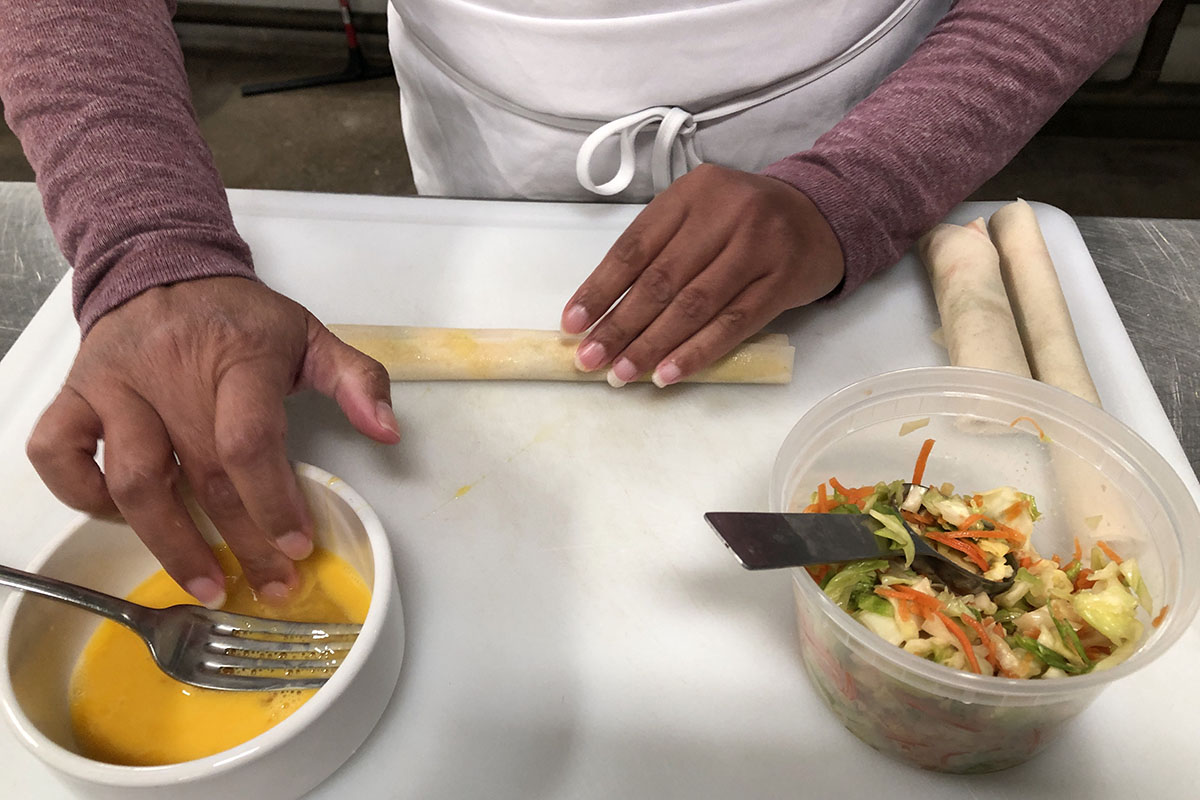 Two hands, one rolling a narrow spring roll, one dipping fingers into a bowl of egg, a bowl of chopped cooked cabbage and carrots next to egg bowl