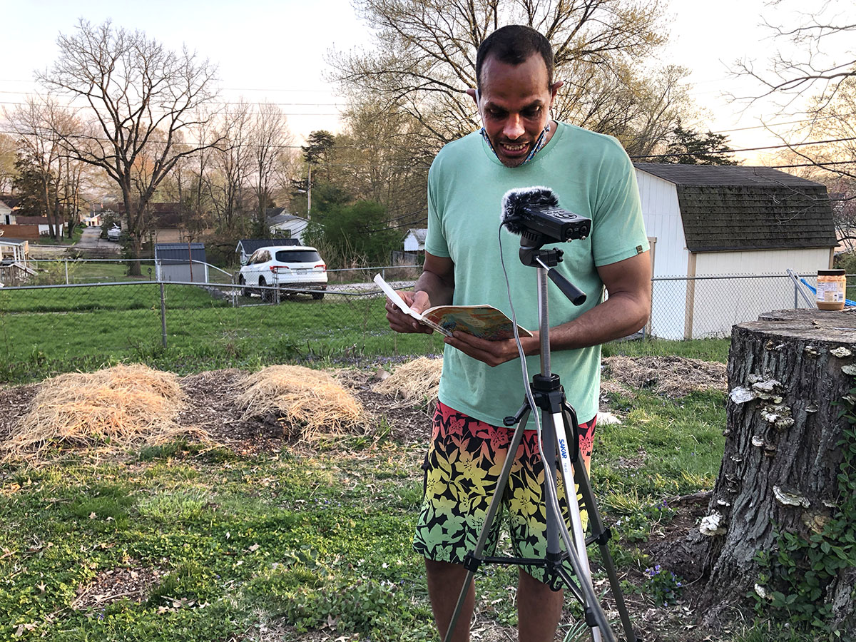 Ross Gay in t-shirt and shorts reading from a book in an early spring garden in front of a microphone on a tripod.