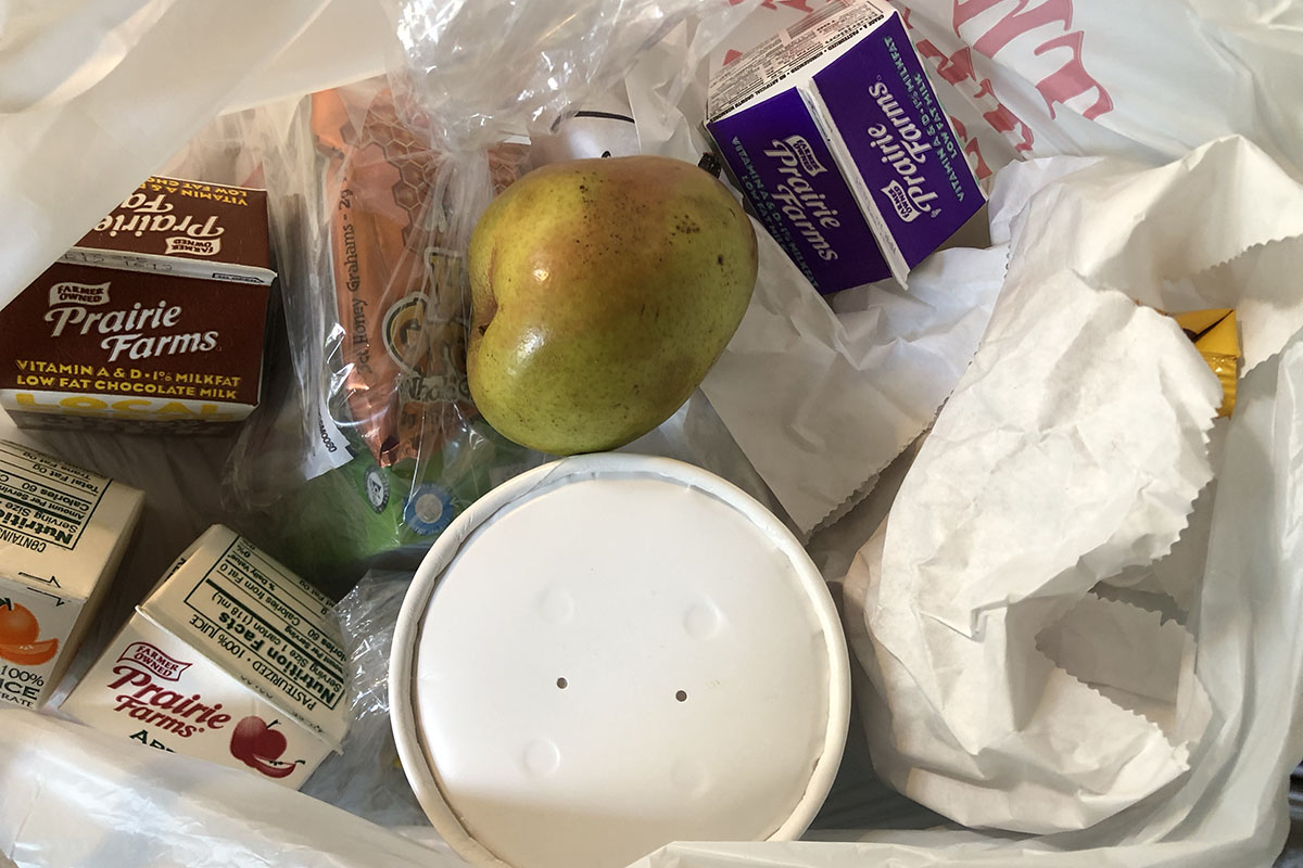 Various food items in a white plastic bag, including small cartons of milk, a pear and a round, white paper container.