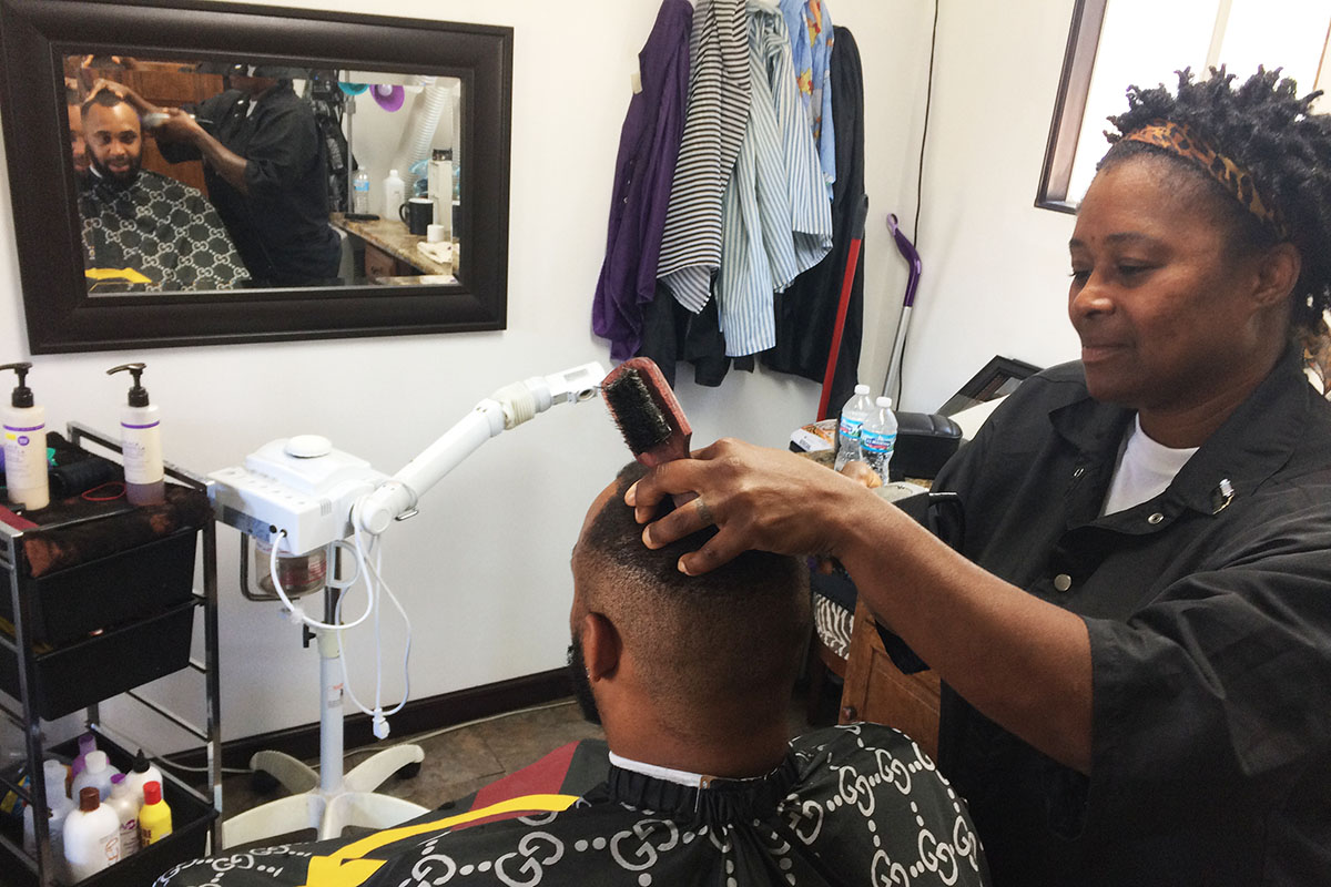 Sharrona Moore in black smock cutting DJ's hair in salon. DJ's face is visible in the mirror