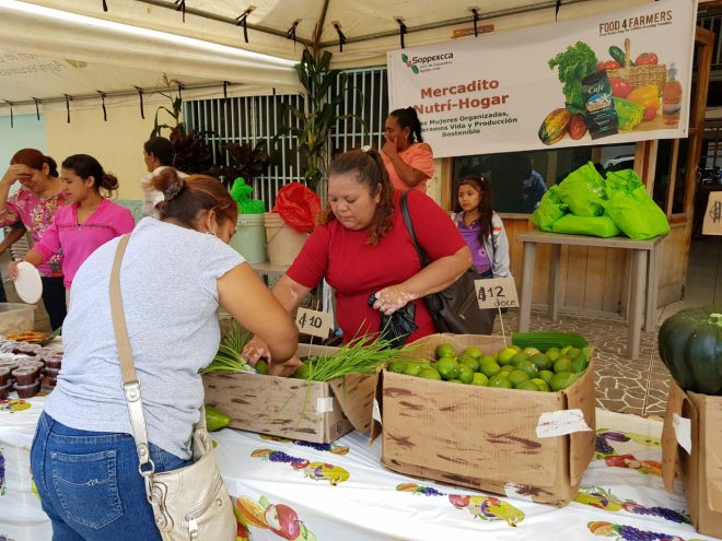 Women at a farmers market with a box of limes on the table and a Spanish language banner in the background