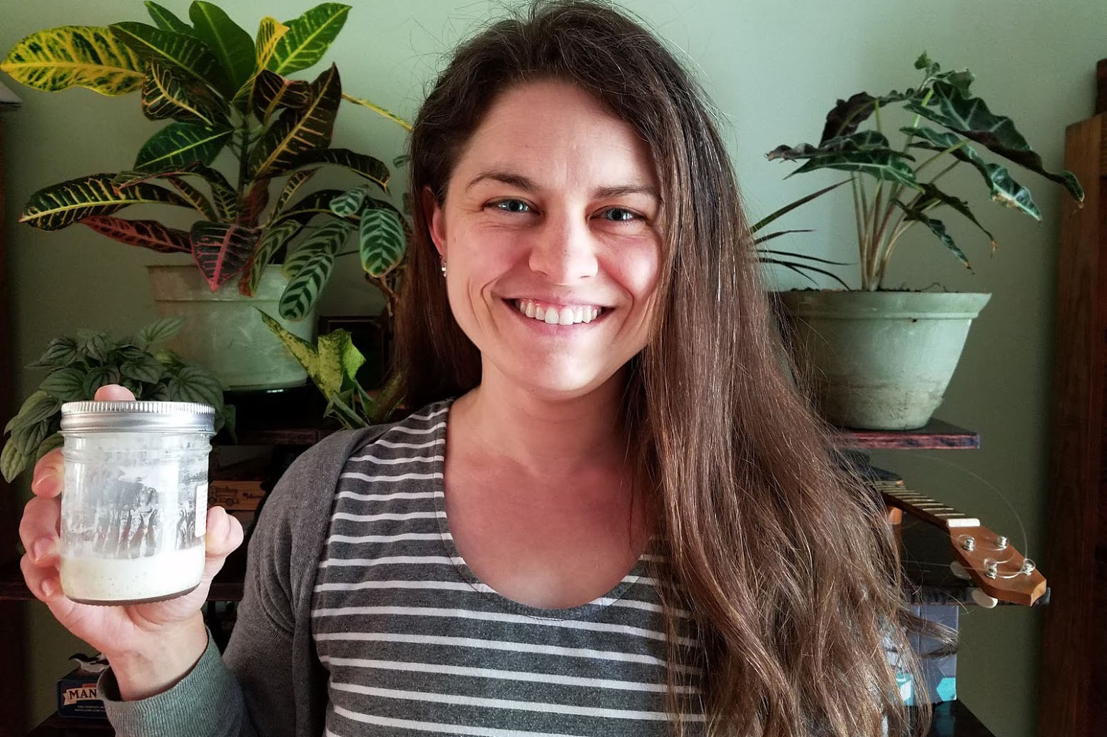 Erin McKenney smiles at the camera and holds up a jar of sourdough starter, a green wall and houseplants in the background.  