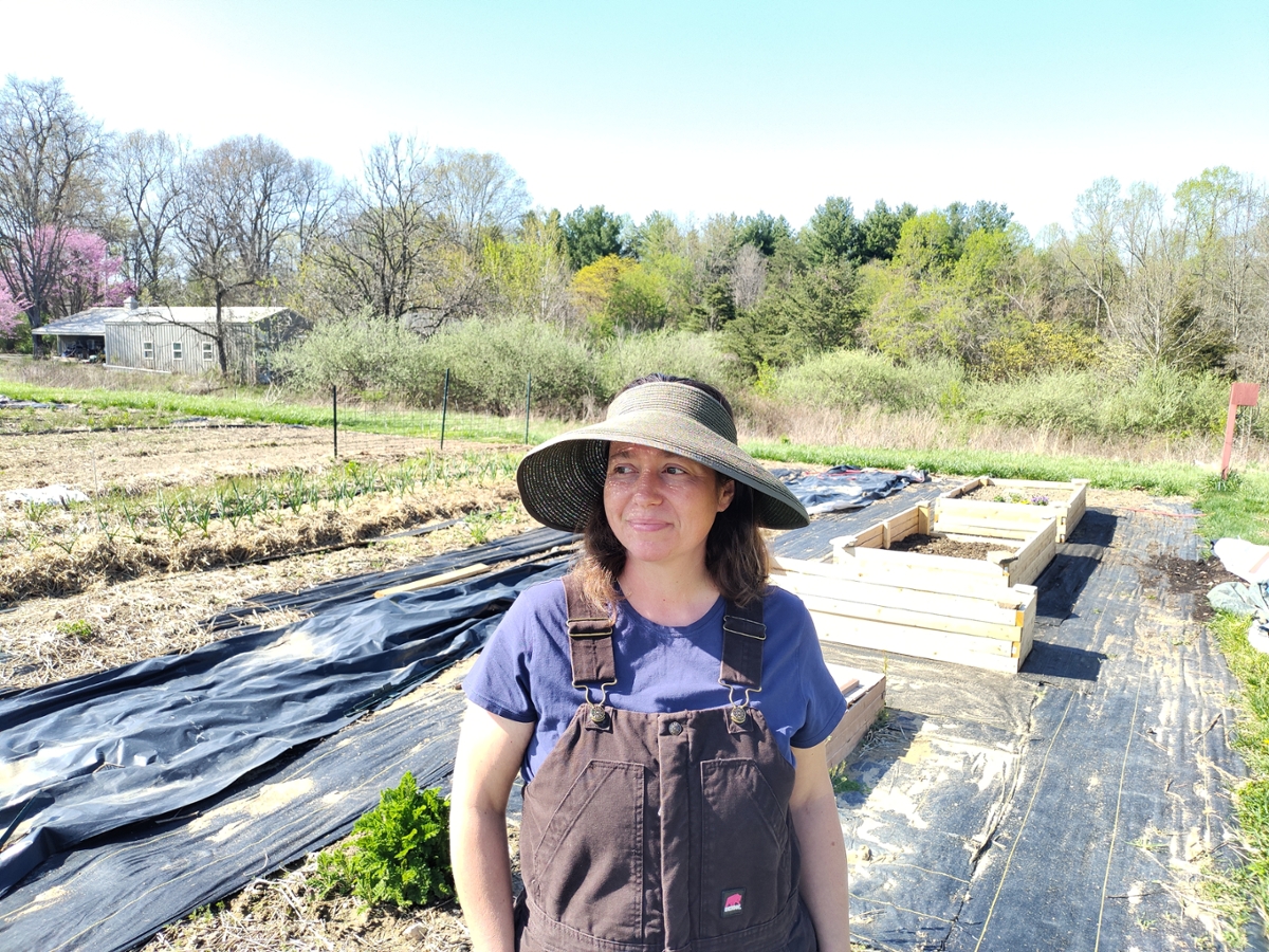A woman Kelli Abdon in a sunhat smiling in front of a small farm field 