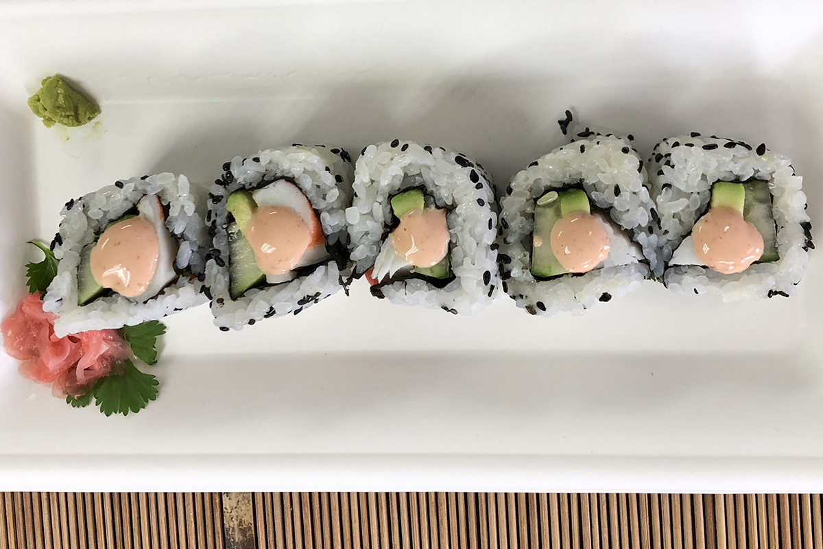 A row of sushi roll segments with a peach colored creamy sauce on each one, aranged on a white plate with a bamboo mat visible underneath.