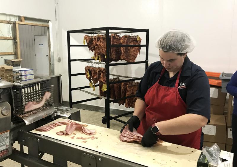 A worker with a red apron, gloves and hairnet, working at a table with ameat slicer. Other cuts of meat hanging on a rack in the background.