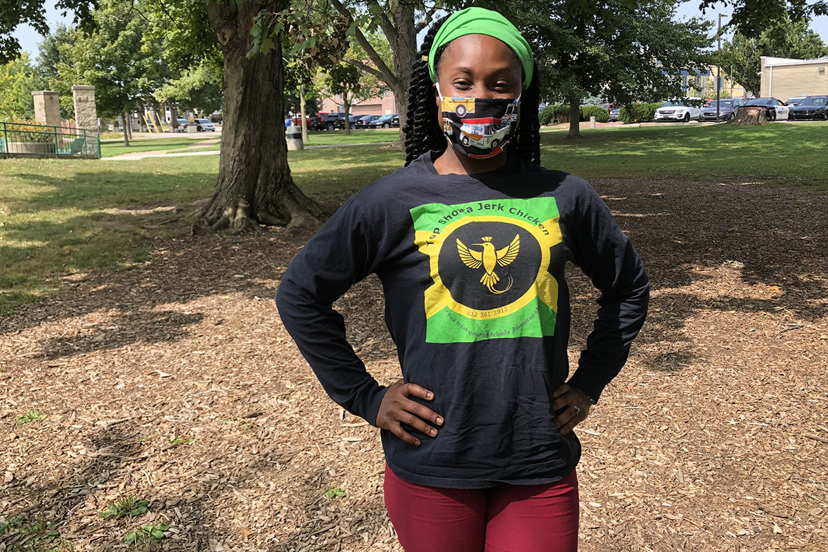 Taneisha Henline in face mask with Top Shotta long sleeved black t-shirt, looking at camera, standing in a park 