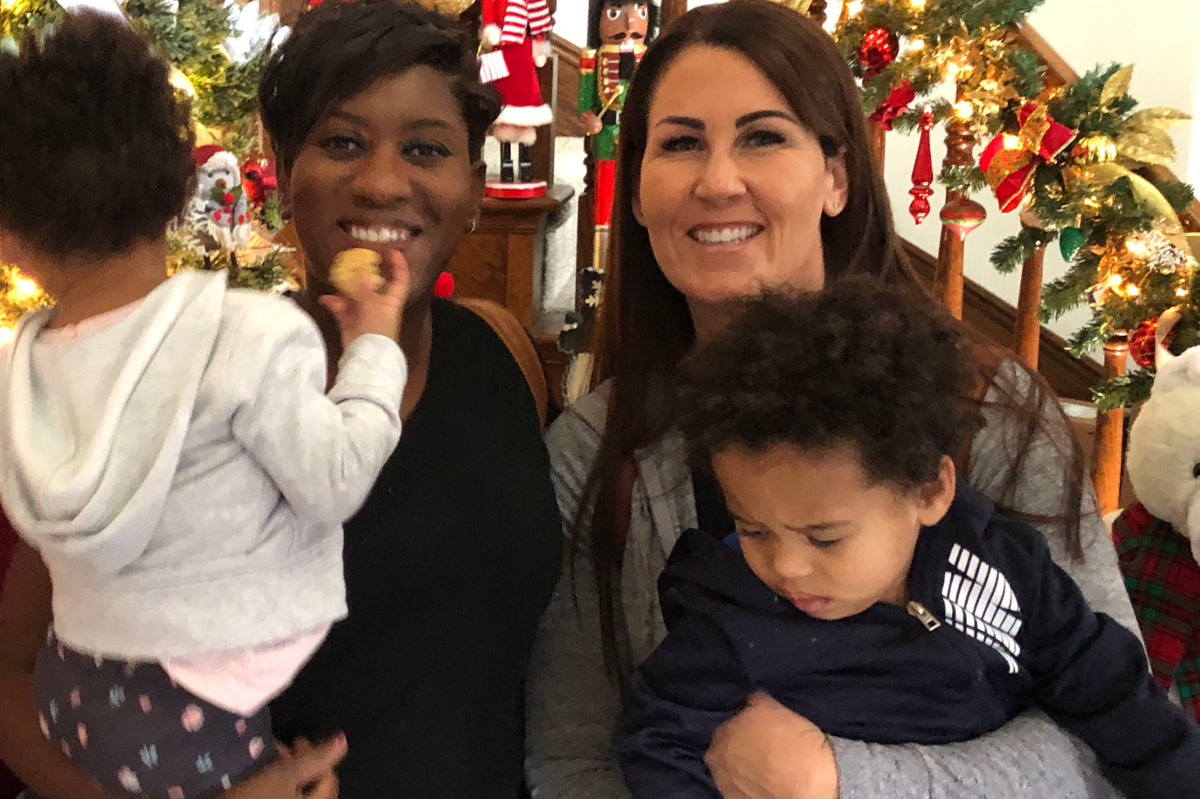 Tiffany-White LaGue and Kristina LaGue smiling at the camera, holding two toddlers with Christmas decor behind them