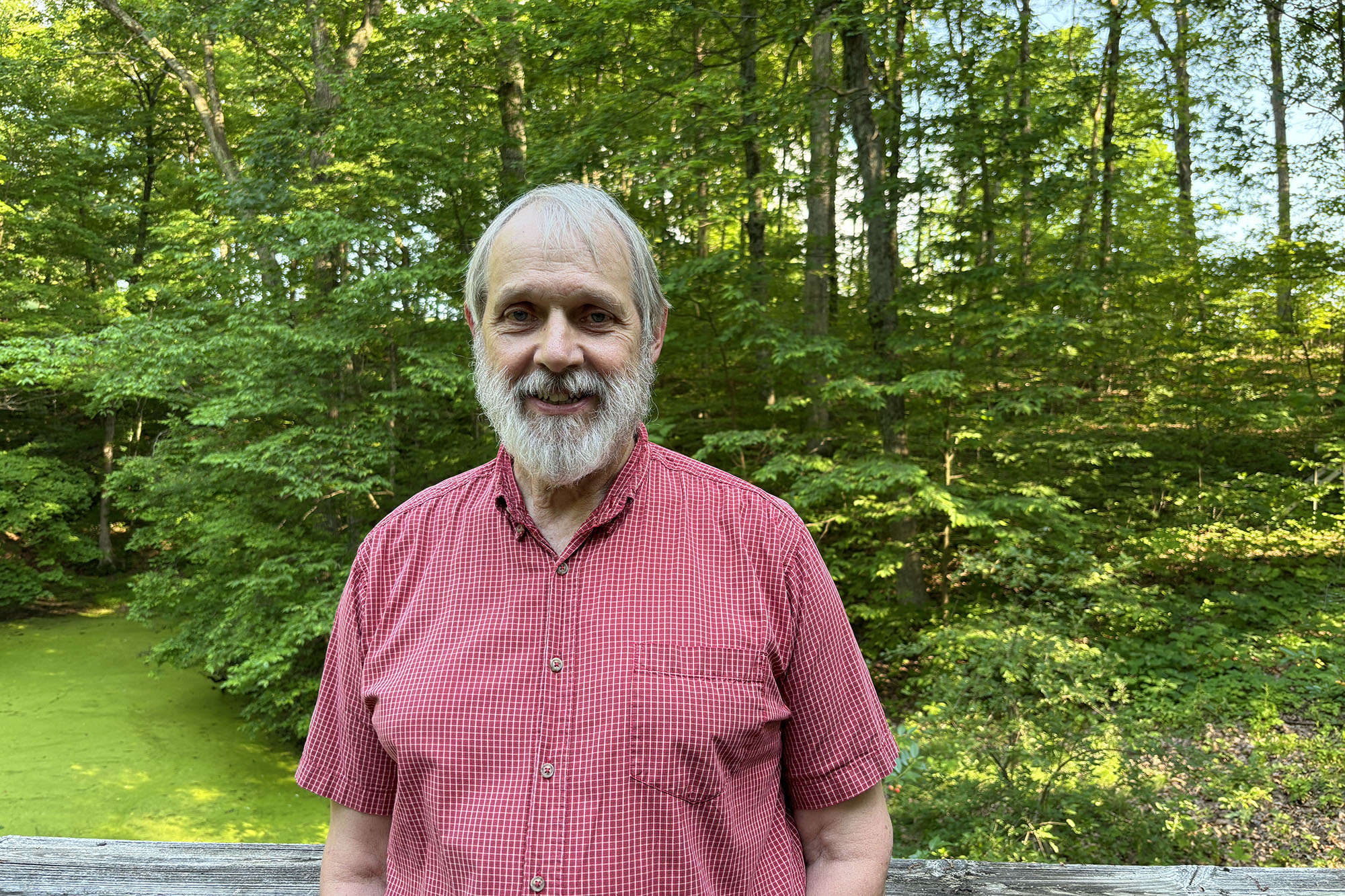 Tracy Branam standing with thick green forest and a green pond behind him