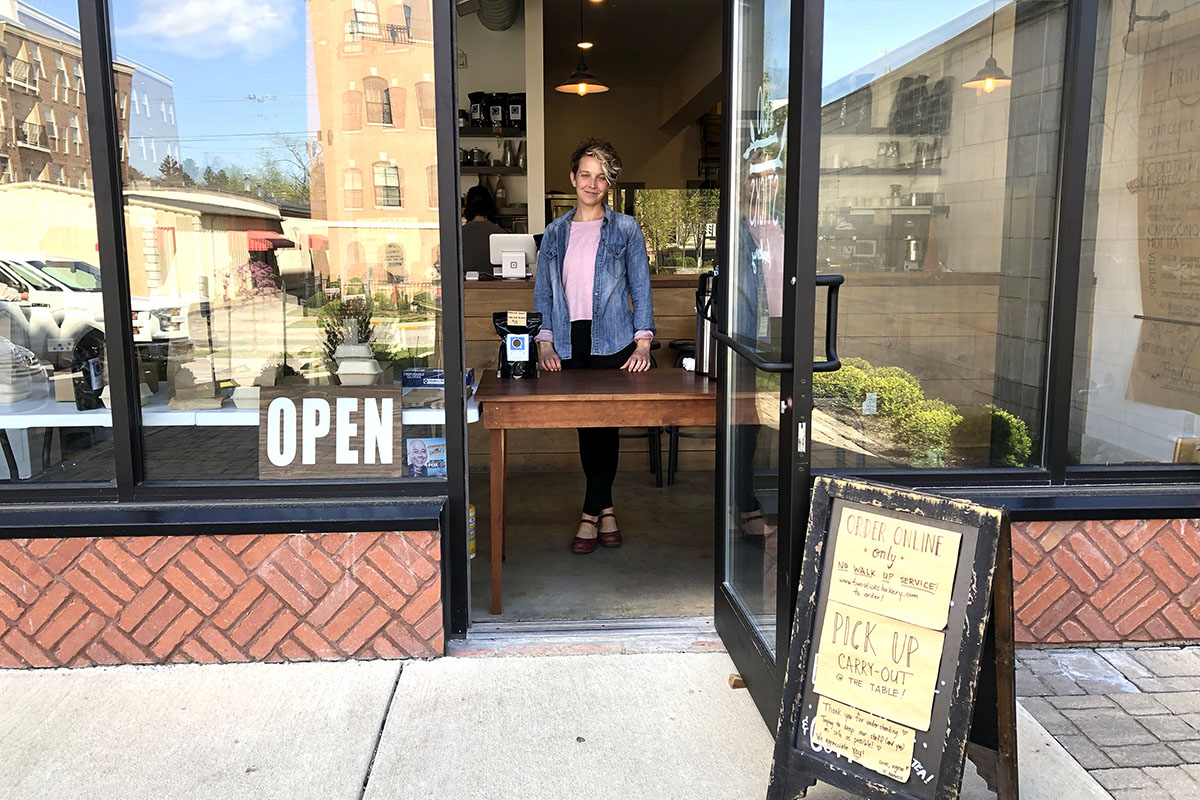 Amanda Armstrong standing behind a table in the open doorway of glass fronted-bakery on a sunny day.