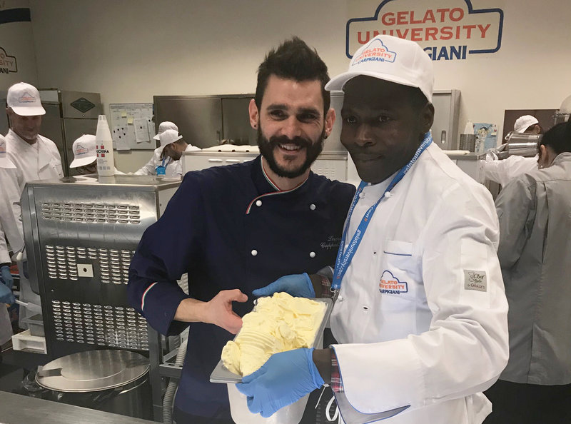 Two guys working in a commercial kitchen holding up a container filled with gelato.
