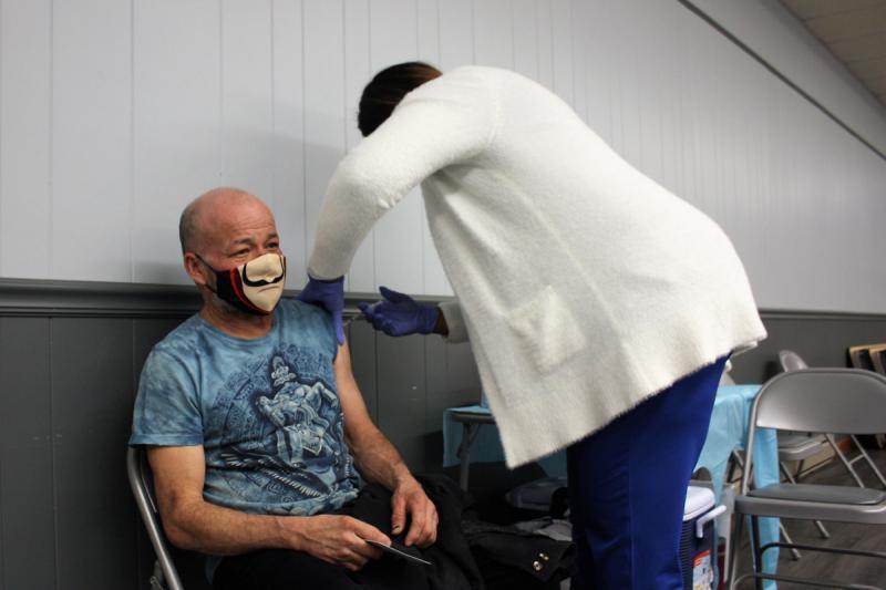 A bald man wearing a humorous mask sitting in a chair while someone leans towards him, administering a shot in his arm.