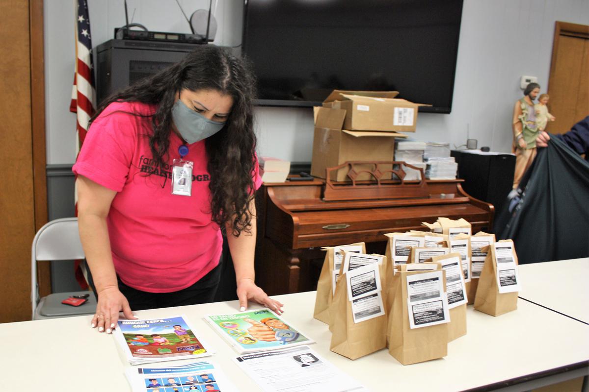 A woman in a face covering in looking down at a table with informational material and brown paper bags with flyers on them. Piano is visible in the background.