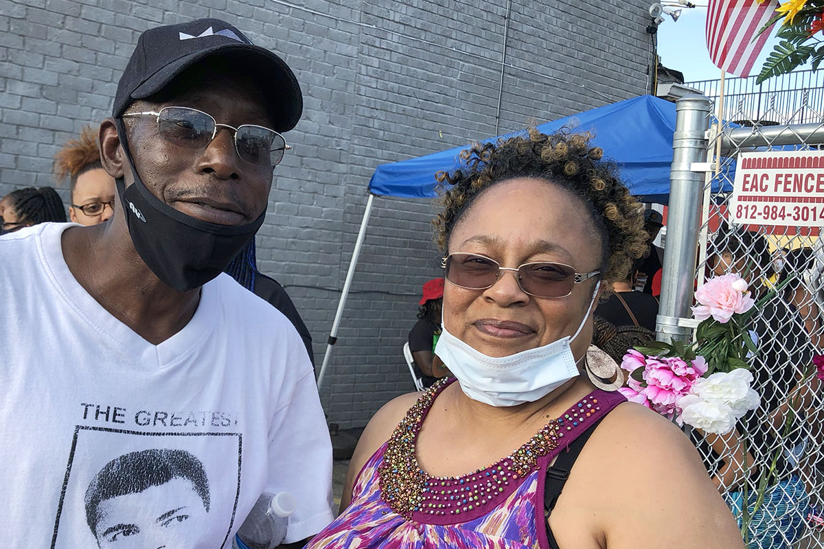 Victor Tellis and Lynnell Blakemore, both with face masks at chin level, with blue tent, a few people and a gray brick wall and flowers on a fence in the background