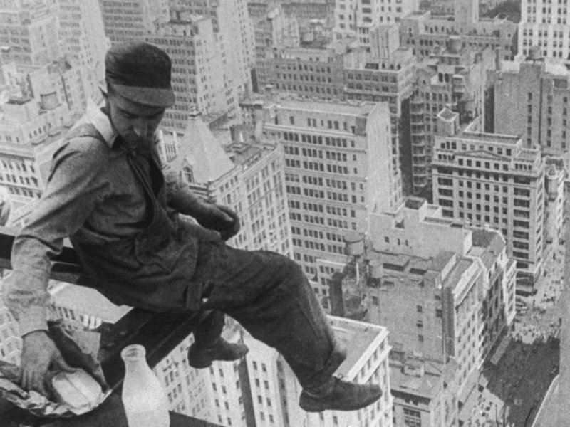 A man in a cap and overalls, seated on the edge of a metal girder, overlooking the city scape, from high above, reaches behind himself for a sandwich. 