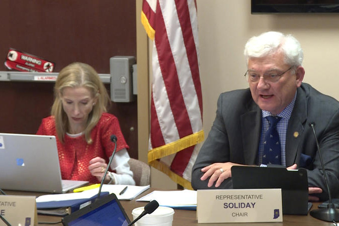 Rep. Ed Soliday sits at a table at the statehouse next to a blonde woman in a red shirt