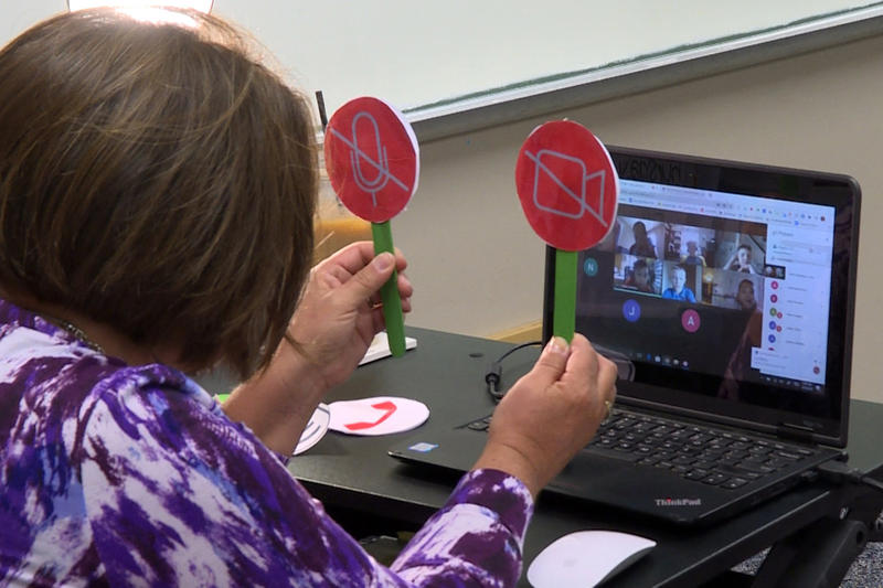 Tami McMahan sits in front of a computer on the first day of school, holding up icons representing the microphone and webcam buttons to help her virtual third grade class see whether they should turn theirs on or off.