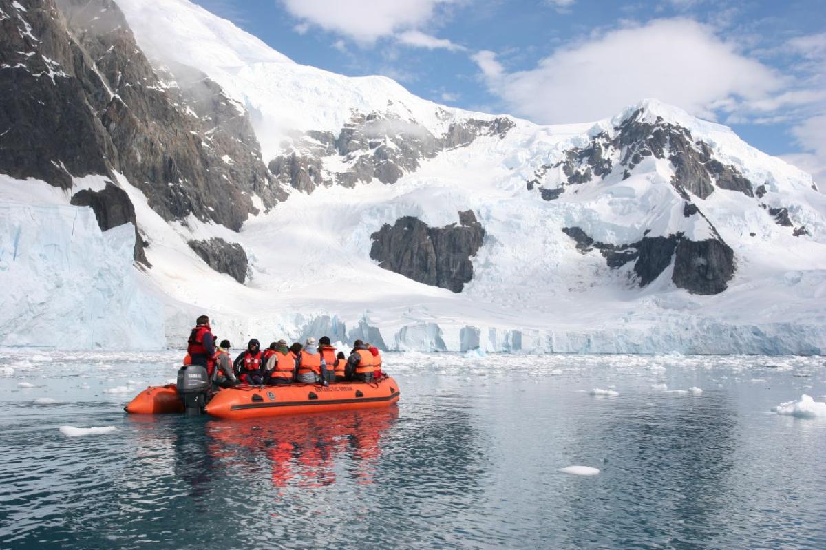 A group wearing orange life jackets sit in a large raft on the water, looking at the large ice formations in Antarctica