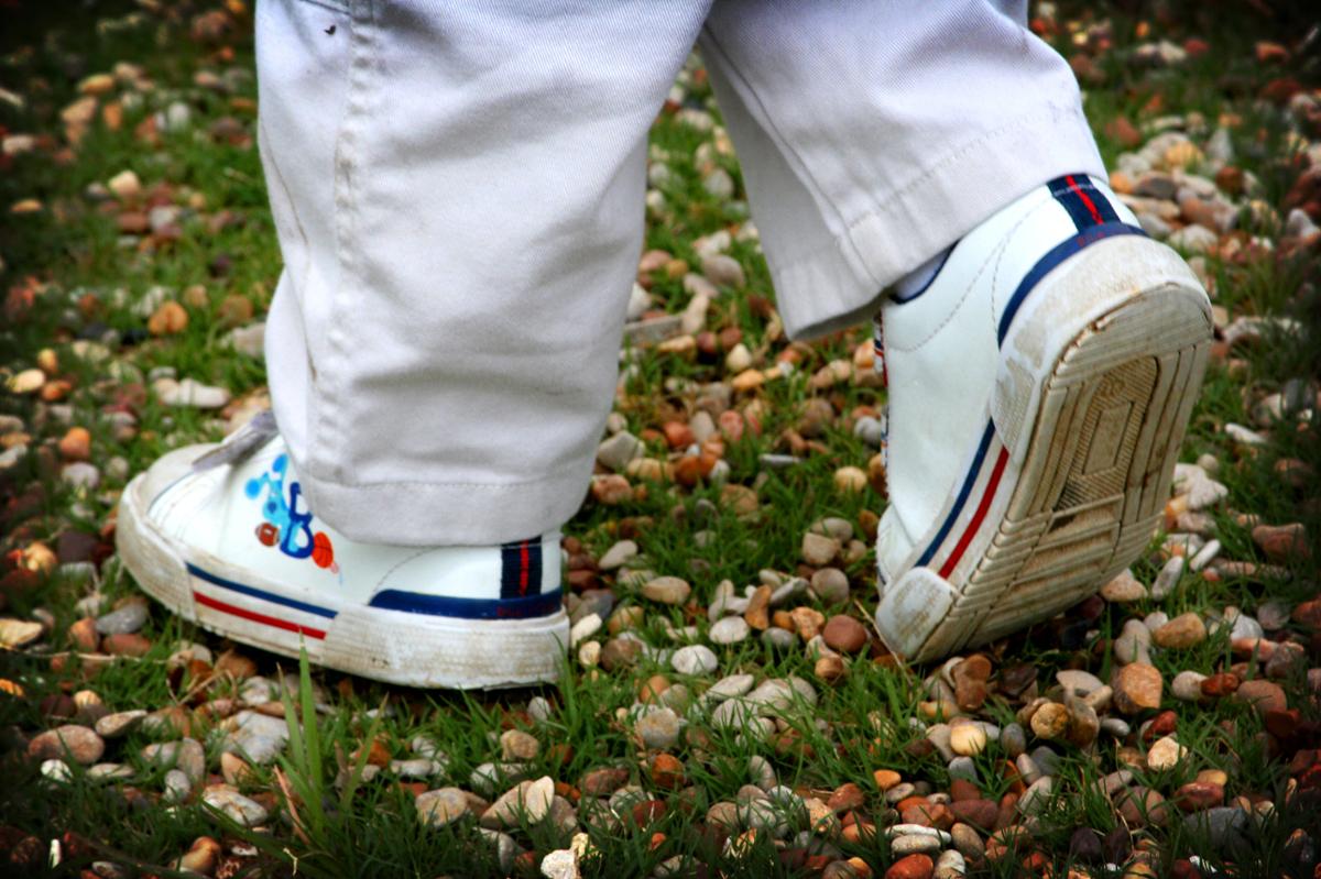 A closeup of a young child's feet with cartoon characters on his white shoes, walking through the grass and leaves