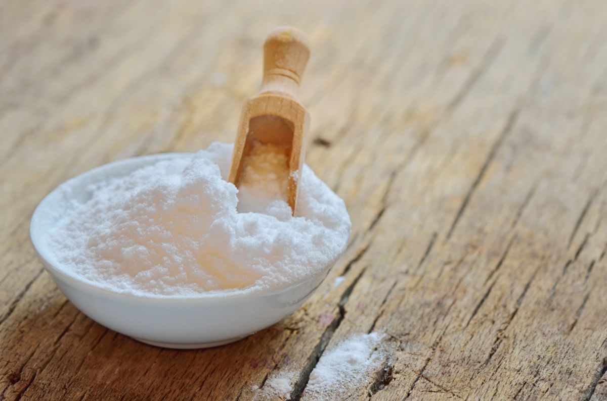 Baking soda in a white bowl with a narrow wooden scoop on a wood surface