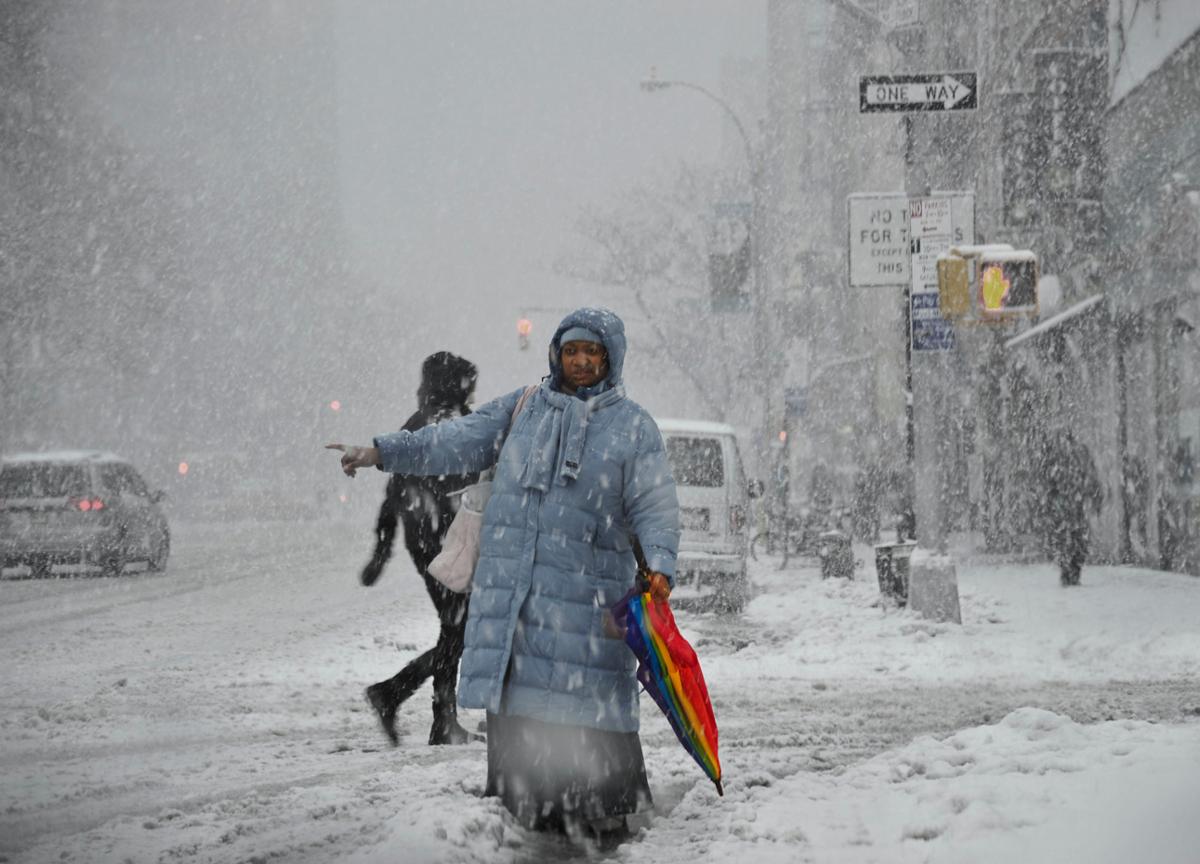 A pedestrian in a long blue coat with a rainbow umbrella stands hailing a cab during a snow storm
