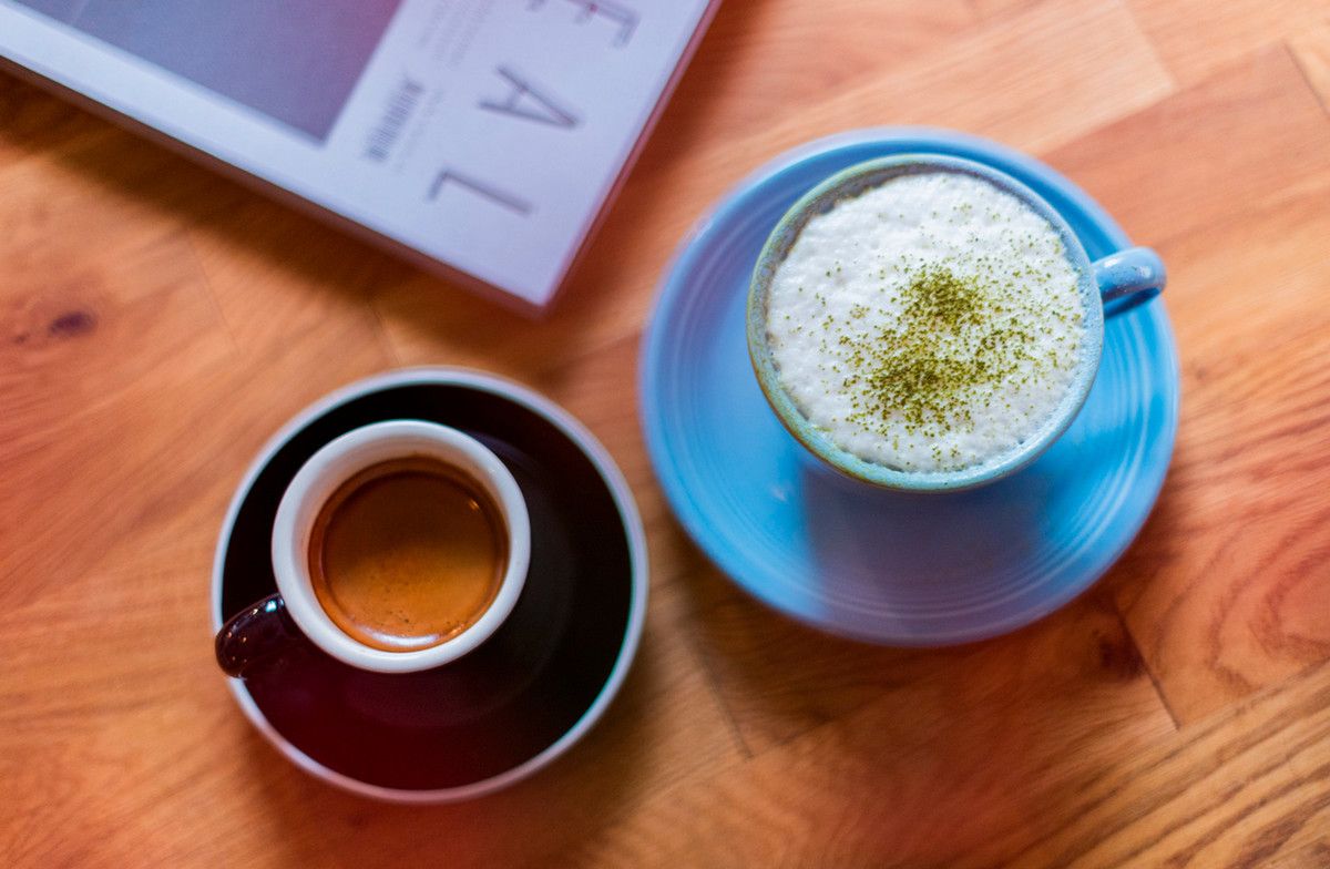 Two mugs on a wooden table as seen from above: a smaller one with a shot of espresso and a larger one full of foamy milk and matcha sprinkled on top