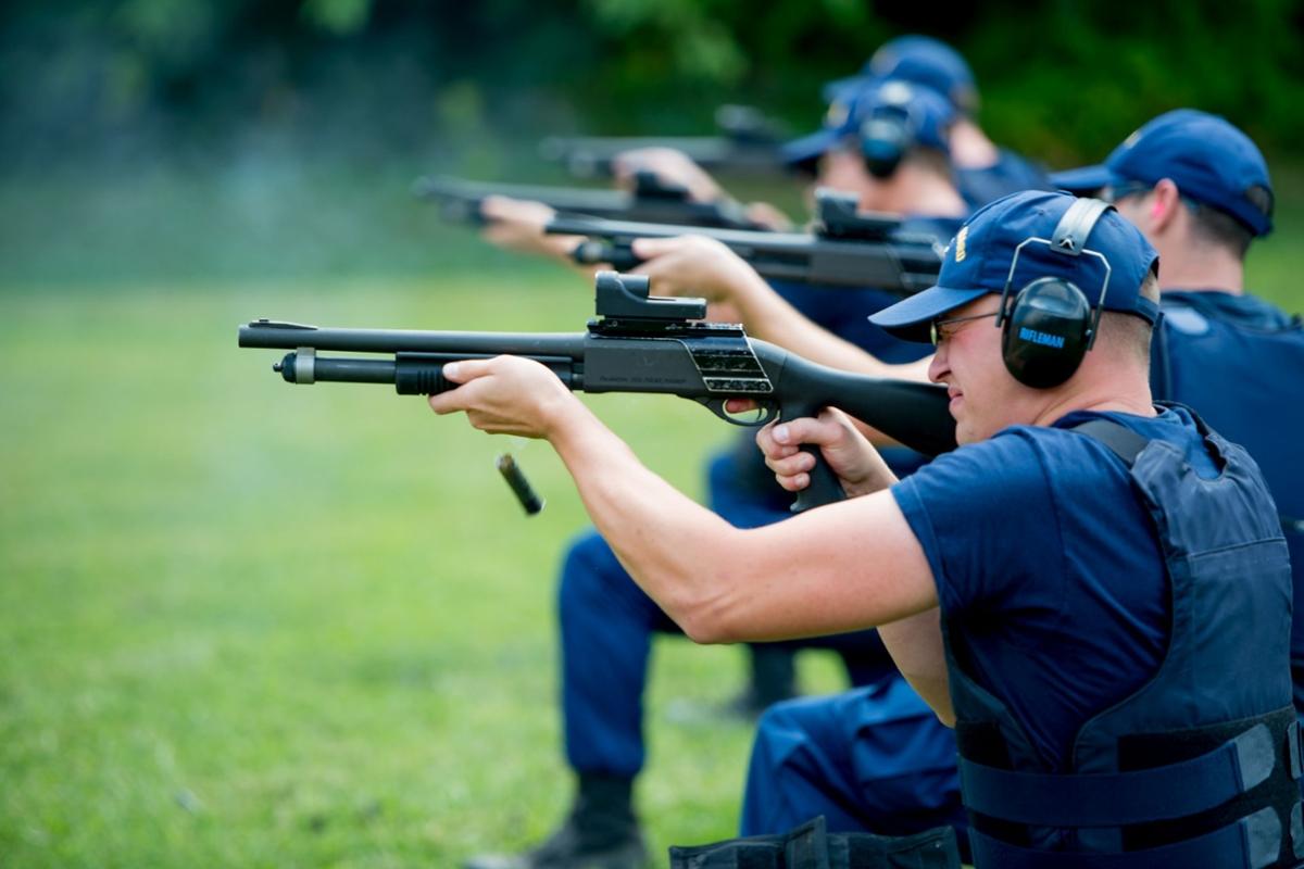 Several members of the Coast Guard dressed in blue and proper gear participate in target practice in a green field