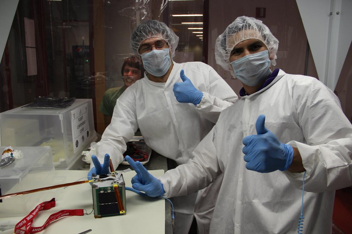 Two scientists give a thumbs up as they pose with a small satellite in the shape of a cube
