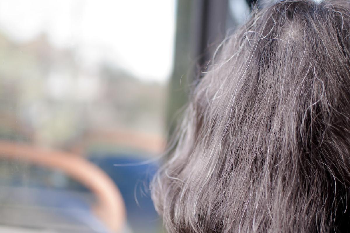 A person with long, grey hair stands with their back to the camera, looking out a window