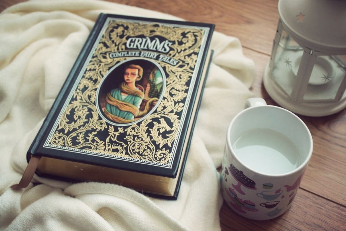 A copy of Grimm's fairy tales on a blanket, on top of a wood table with a mug and lantern sitting nearby