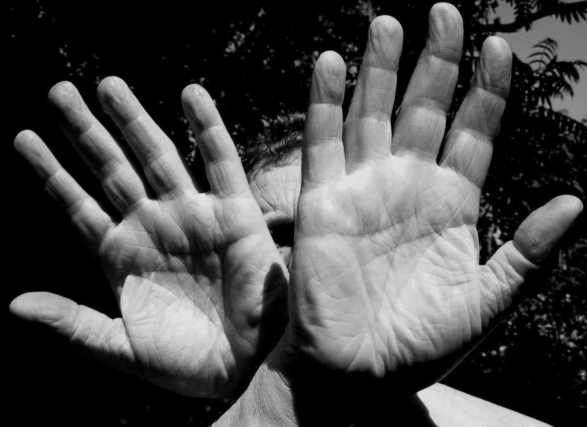 A man holds the palms of his hands up to the camera blocking his face, in black and white
